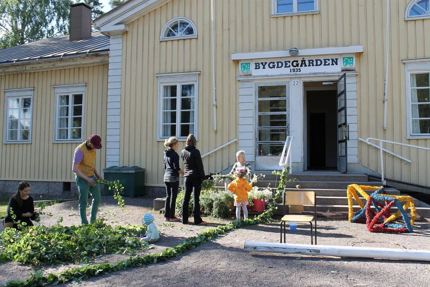 Ett stort gult trähus med skylten Bygdegården ovanför dörren. Utanför huset står människor och sköter lövningen och bindningen av midsommarstången i Tenala.