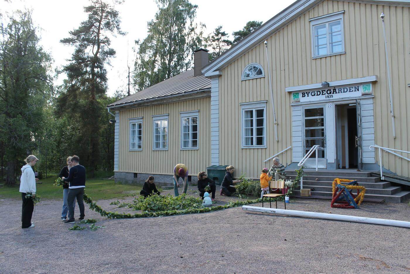 Människor står utanför ett stort gult trähus. De håller på att fixa lövningen och bindningen av blommor till Tenalas midsommarstång.