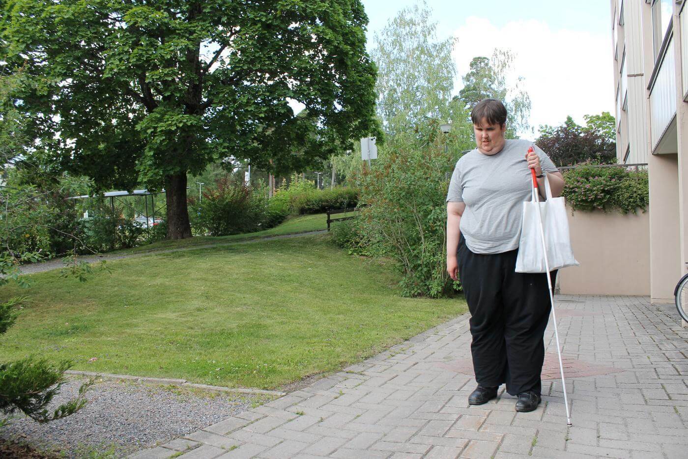 En kvinna står med en vit tygpåse och vita käppen i ena handen. I bakgrunden syns en gräsmatta och gröna träd.