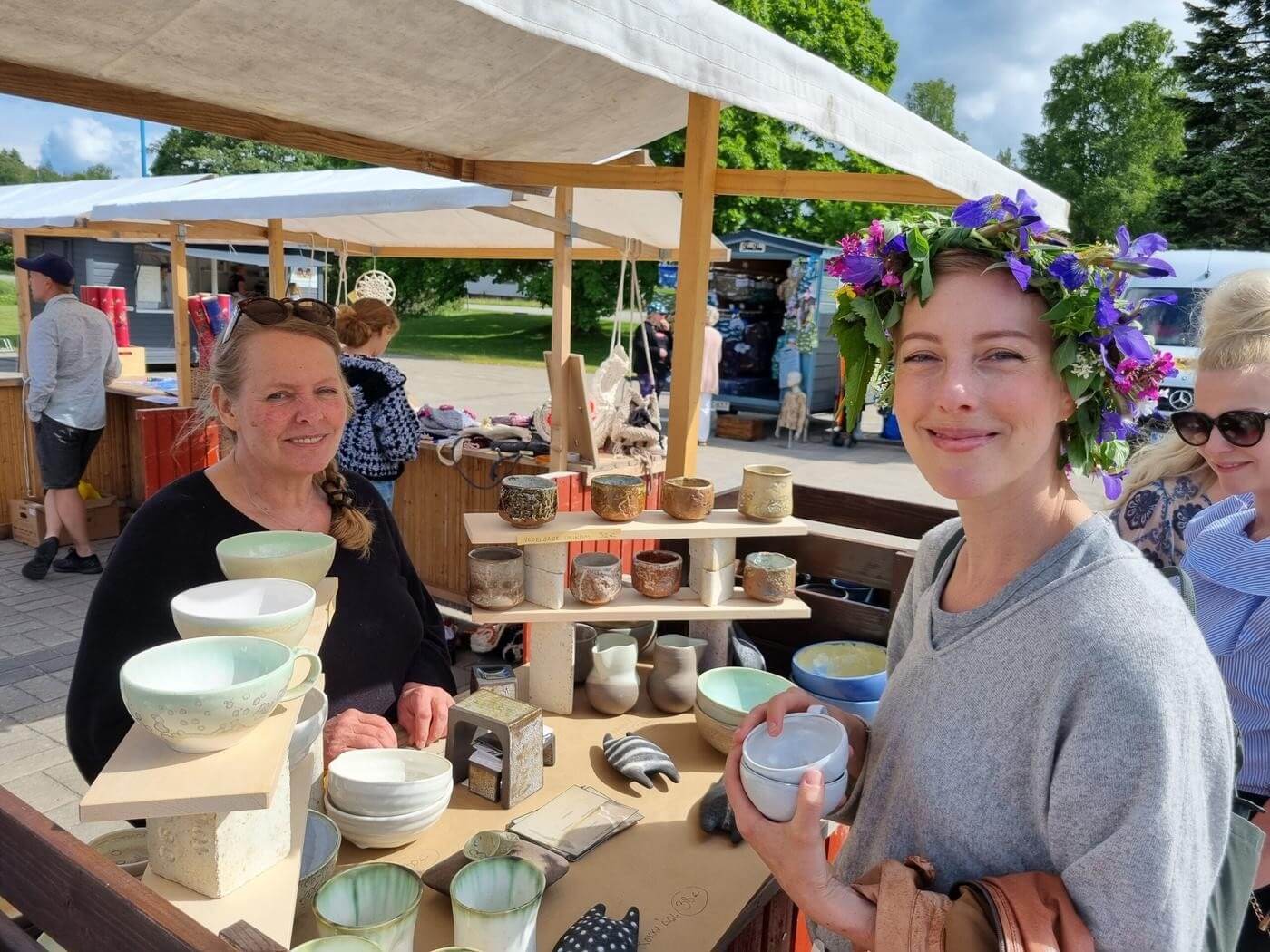 Två kvinnor på sommarmarknad. Ena kvinnan har en sommarkrans på huvudet.