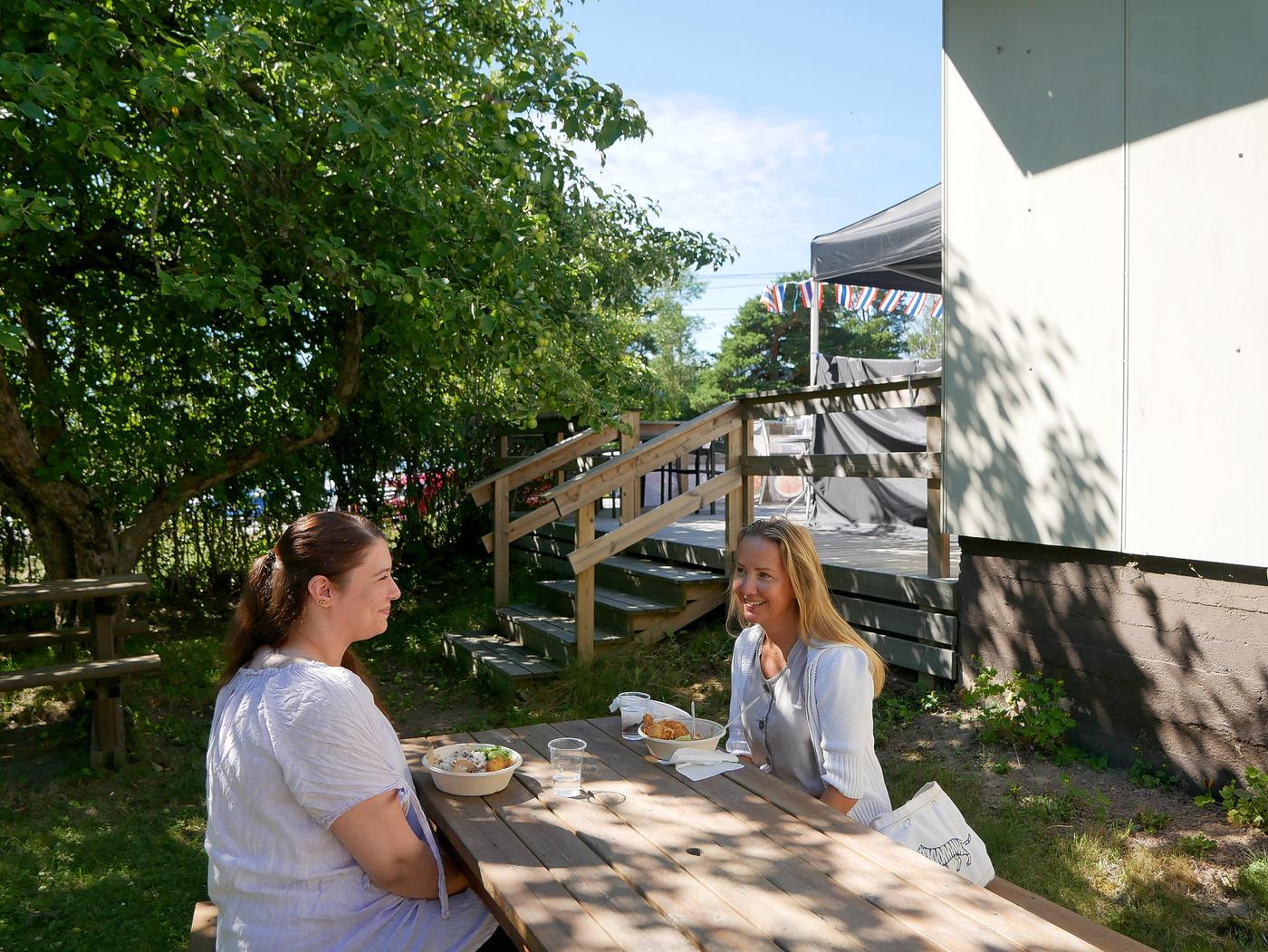 Två kvinnor lunchar i trädgård.