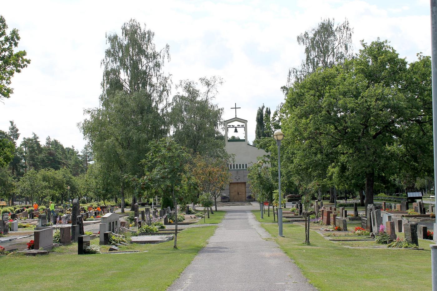 Bild på nya gravgården i Ekenäs. Gröna träd och en liten grusgång mot ett av kyrkans hus på gravgården. Bredvid  grusgången finns flera gravstenar.