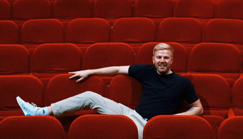 En ung kille som gör standup sitter i röda bänkar på en läktare inomhus.