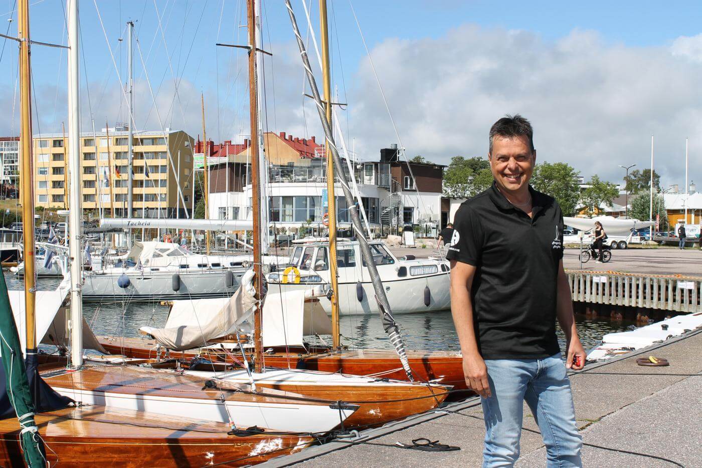 En man står utomhus i östra hamnen i Hangö. Mannen tittar in i kameran och ler. i bakgrunden ser man flera båtar.