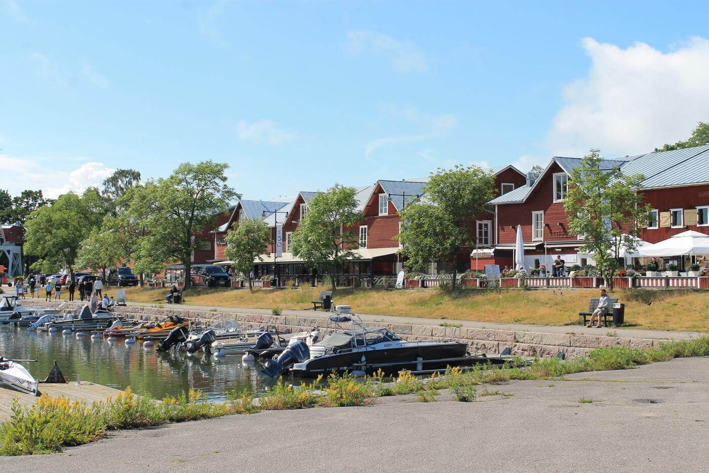 Vatten och några båtar som står vid bryggan i Östra hamnen i Hangö. röda trähus syns längs med gatan som går längs med vattnet. Många människor sitter på husens terrasser.