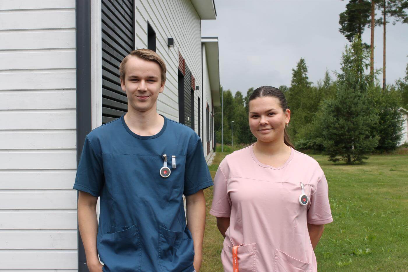 En ung man står bredvid en ung kvinna, de tittar in i kameran och ler. Både två är klädda som vårdare då de sommarjobbar på ett äldreboende. I bakgrunden ser man gröna träd och ett en vägg av ett grått trähus.