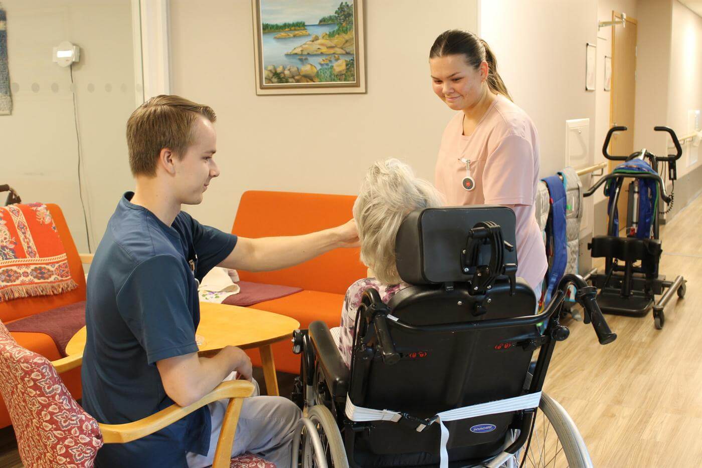 En ung man sitter på en stol och matar en klient som sitter i rullstol. Man ser endast rullstolens baksida och bakhuvudet på klienten. En ung kvinna står bredvid klienten och ler.