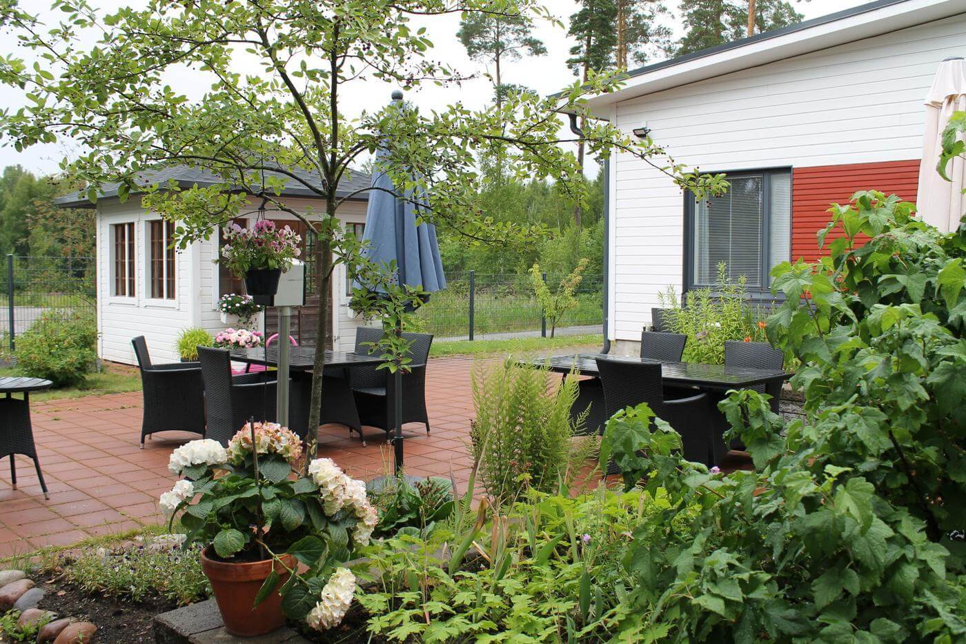 En trädgård med flera blommor och några träd. Några utemöbler är placerade i trädgården och även ett litet vitt lusthus finns på palts i trädgården. I högra hörnet ser man en liten del av ett vitt trähus. Huset man skymtar är äldreboendet Hagahemmet i Ekenäs.