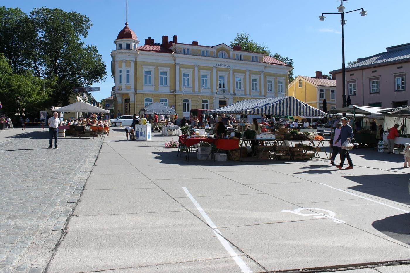 En solig torgdag i Ekenäs. Många människor står vid torgstånden. I bakgrunden ser man Kungsgatan och det gamla stadshuset.