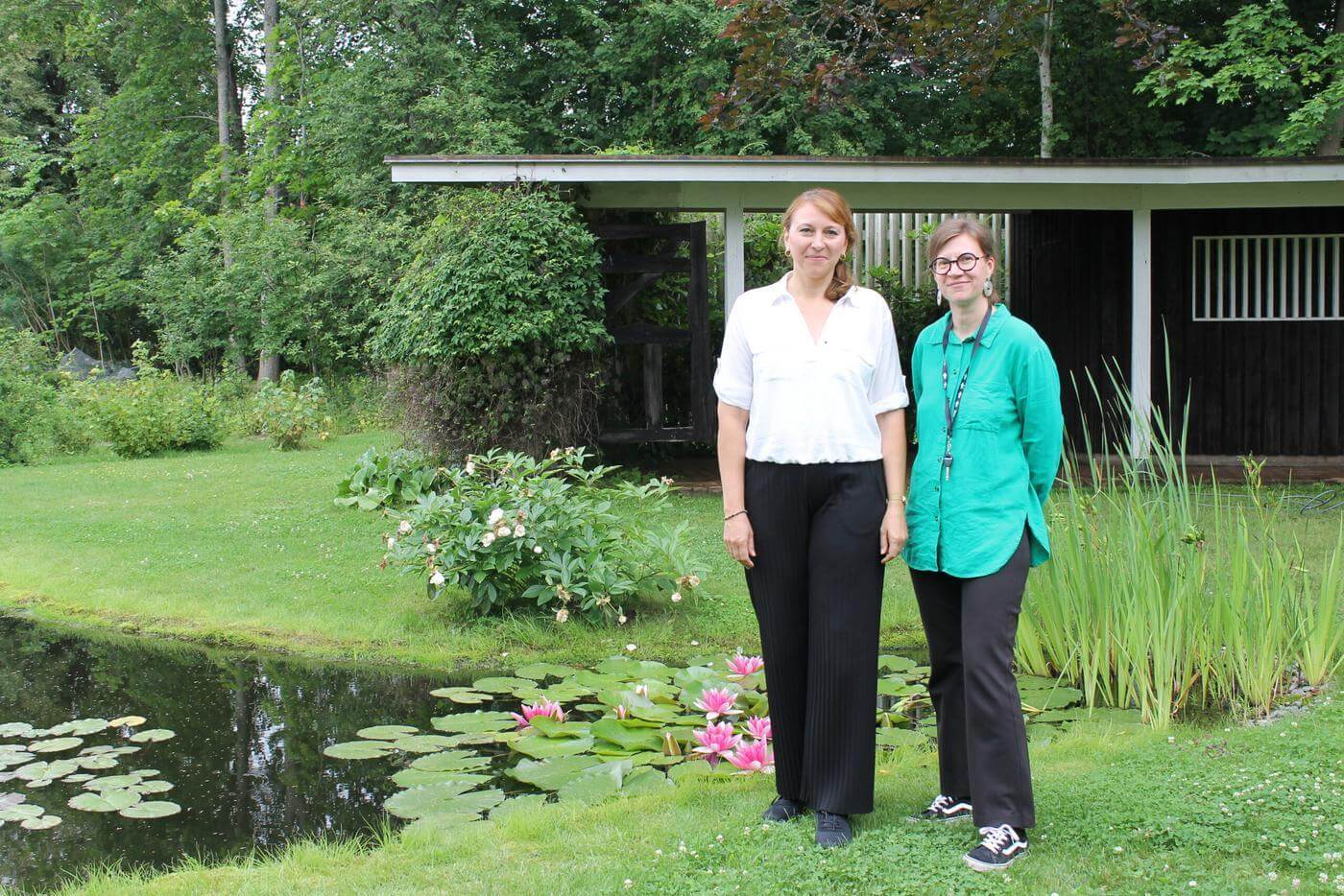 Två kvinnor står bredvid varandra utomhus i en trädgård med gröna träd och buskar. Kvinnorna kommer att arrangera en finländsk-tysk kulturfestival i Ekenäs.