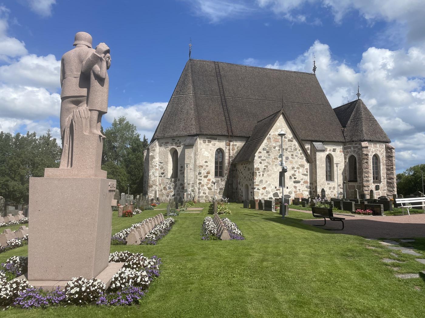 Bild på en gråstenskyrka med hjältegravar och vackra blommor. 