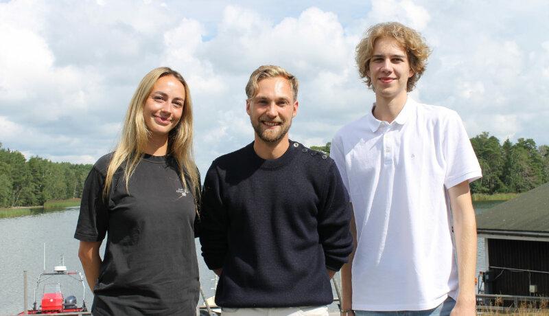 En ung kvinna och två unga män står bredvid varandra och tittar in i kameran. De deltar i ett seminarium om Östersjön i Tvärminne. I bakgrunden ser man vatten och en mulen himmel.