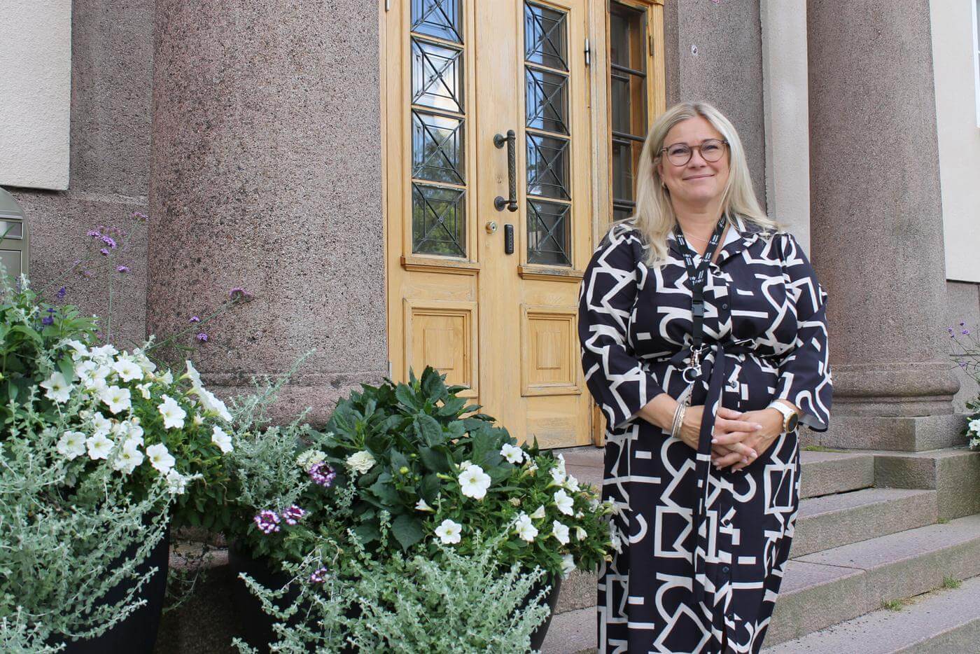 En kvinna står framför stadshuset i Ekenäs. Hon är stadens ny undervisningschef. Bredvid henne finns tre stycken större blomkrukor med blommor i.