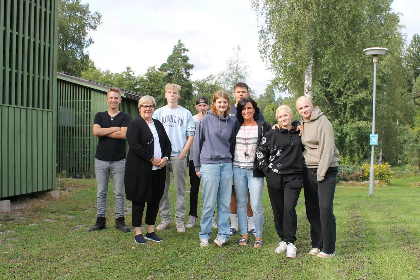 En grupp unga studerande vid Axxell står utomhus på en gräsmatta tillsammans med Axxells enhetschef Beatrice Westerlund och internathandledaren Bettina Lindholm.