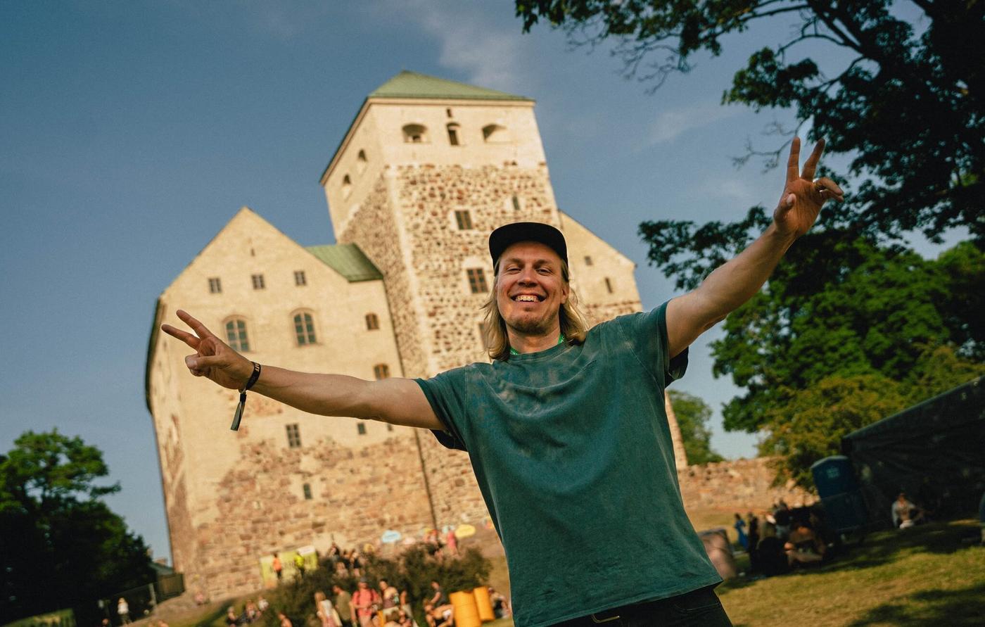 Leende man som håller upp båda händerna och visar "peace"-tecknet. Bakom honom syns Åbo slott.